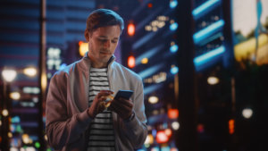 Man Using Smartphone Walking Through Night City Full of Neon Colors and Entertainment. Stylish Young Man Using Mobile Phone, Posting on Social Media, Online Shopping, Texting on Dating App